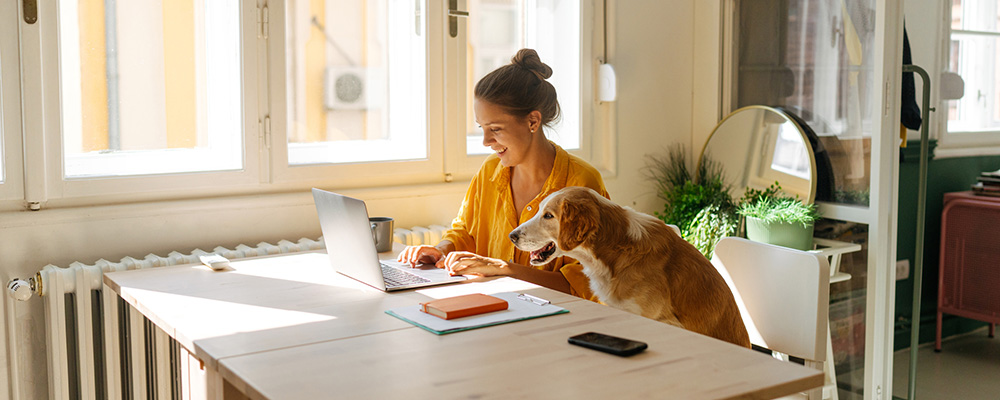 Woman working on laptop at table with dog sitting beside her.